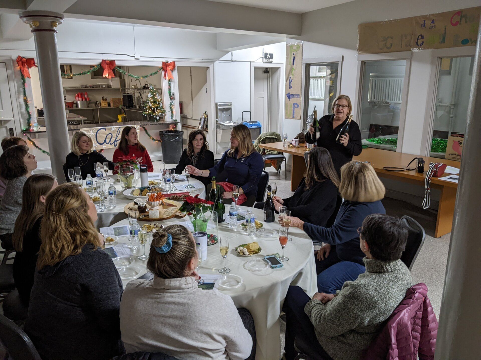 Group of people gathered around a table; a woman speaks, holding a bottle. Festive decorations are visible.