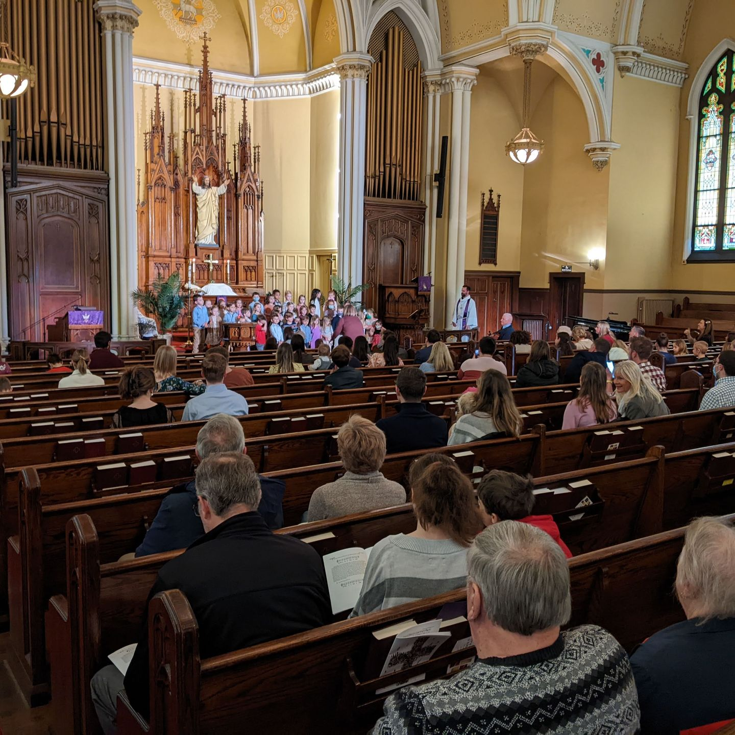 Choir singing at the altar in a church. People in pews are listening. Light wood, ornate details.