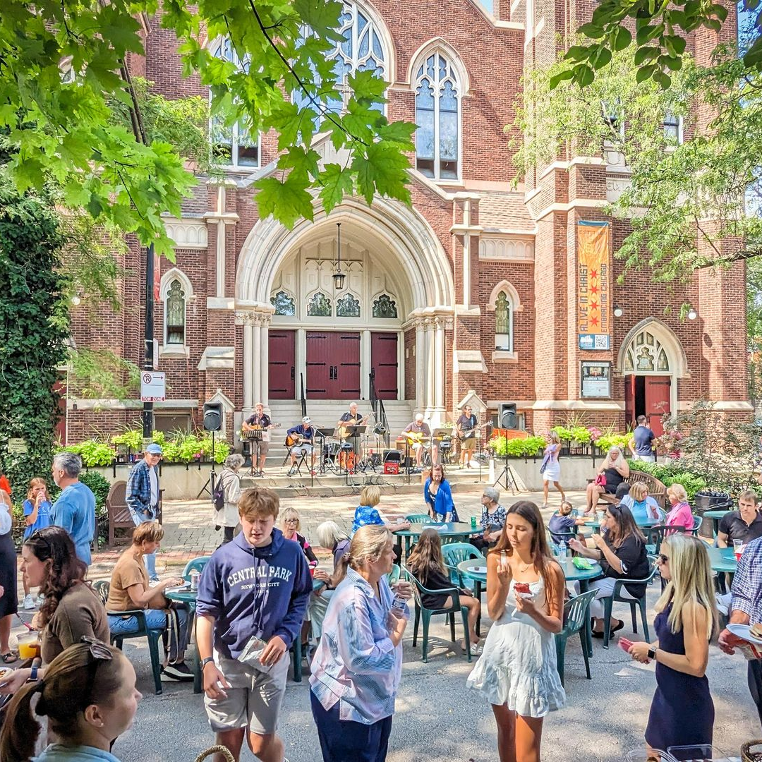 Outdoor event: People gather, eat, and listen to a band in front of a brick church. Green trees overhead.