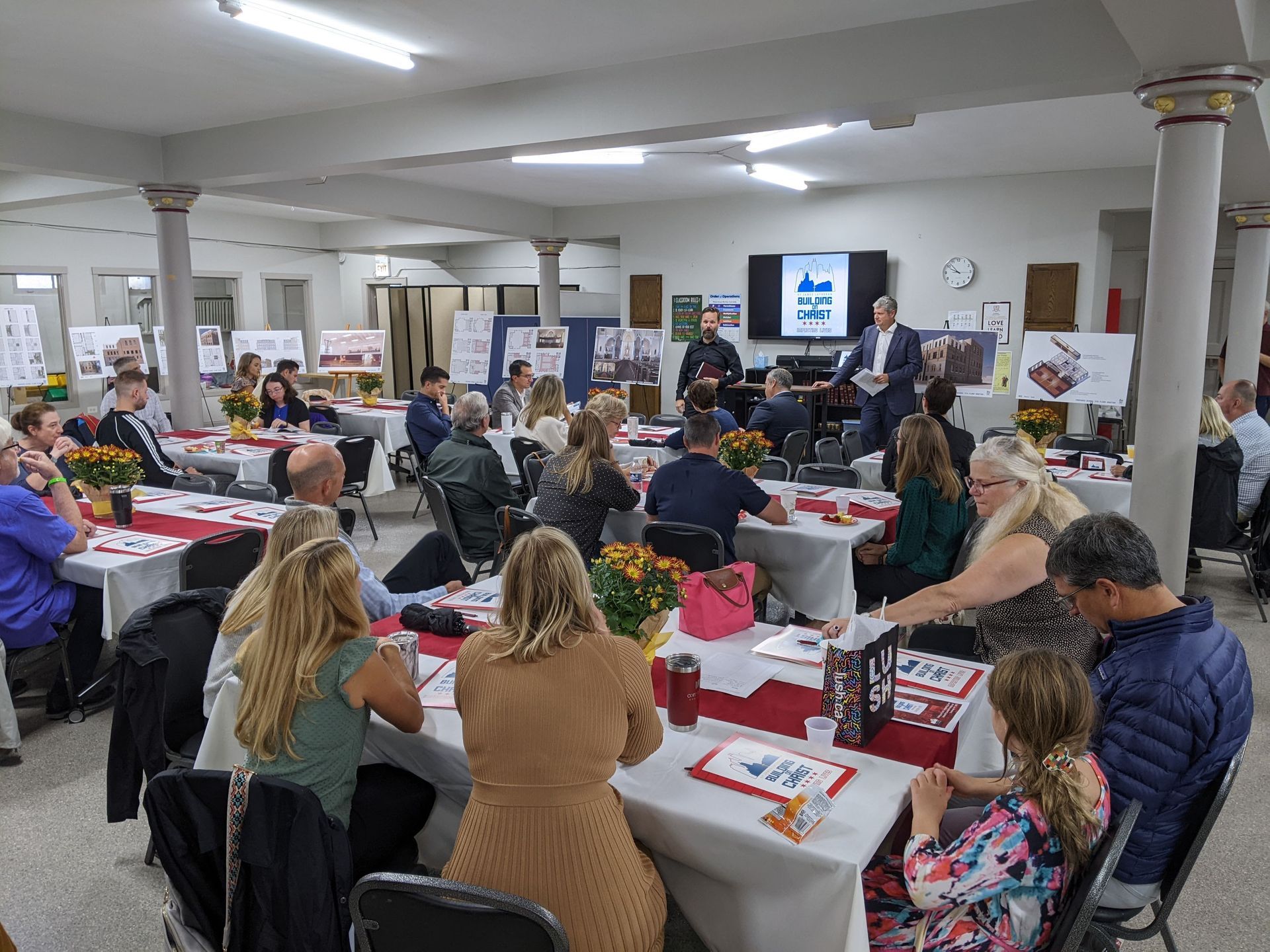 People seated at tables in a hall with a speaker at the front giving a presentation.