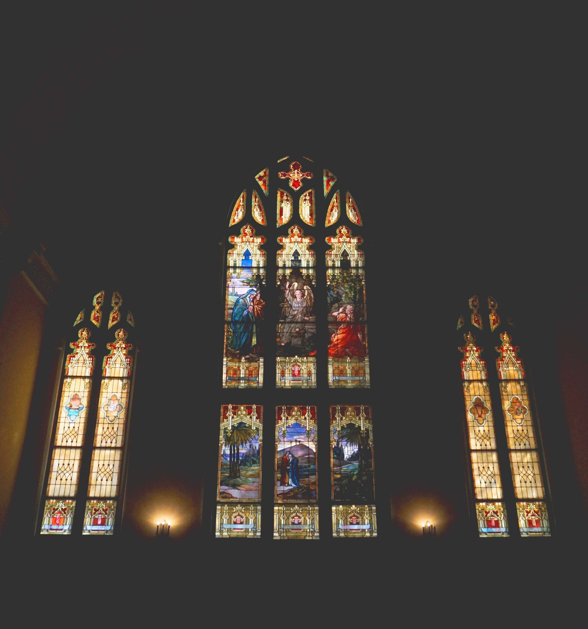 Stained glass windows in a dark room. Central window is largest, with detailed colorful imagery.