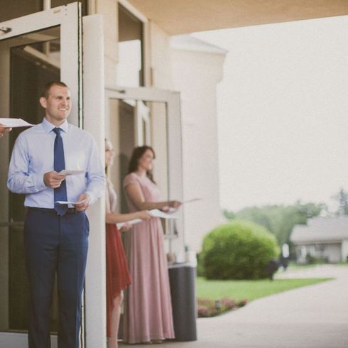 Two people stand near a building entrance holding papers, with a walkway and greenery in the background.
