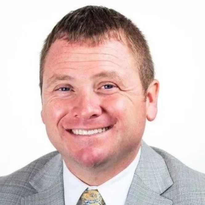 Smiling man in a light gray suit and patterned tie against a white background