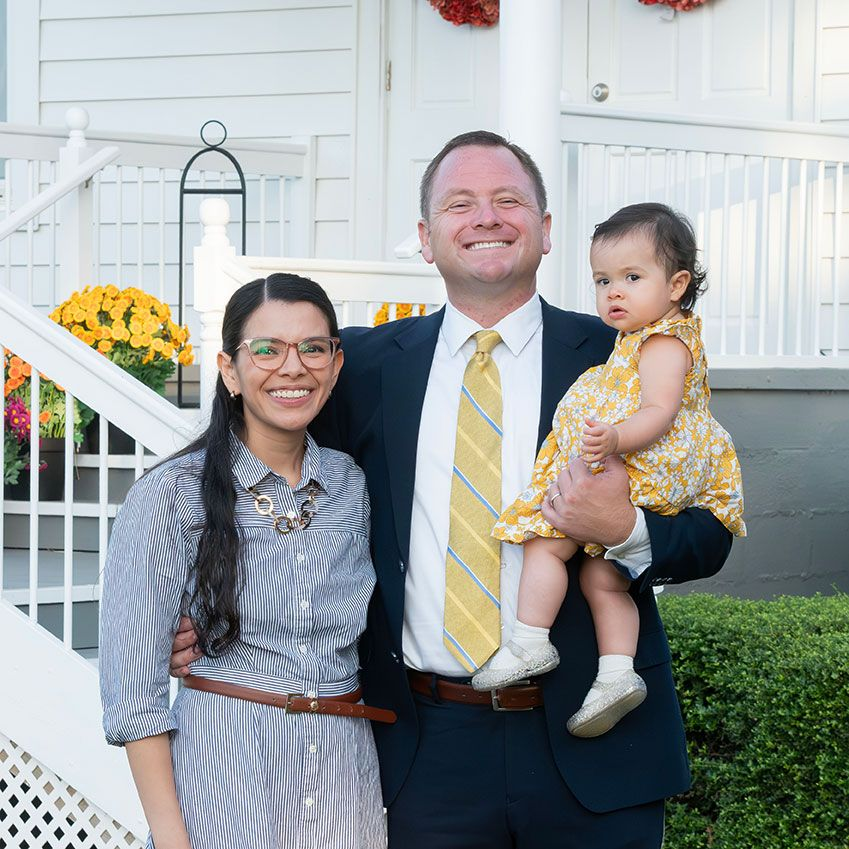 Three people posing on a porch, smiling, with a white house and yellow flowers behind them