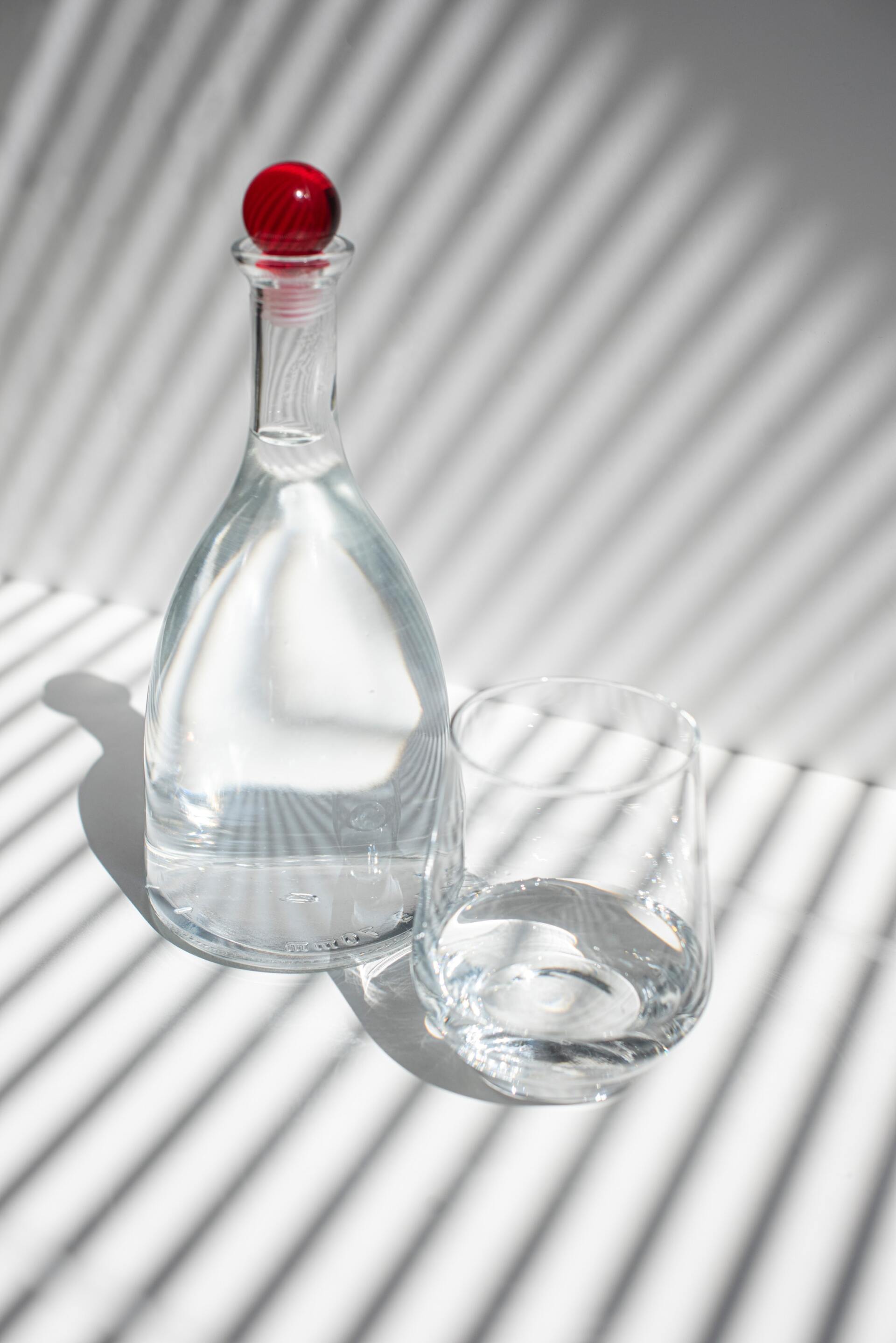 Une bouteille d'eau en verre transparent avec un bouchon rouge et un verre d'eau assorti, sur une surface blanche ombrée de rayures.