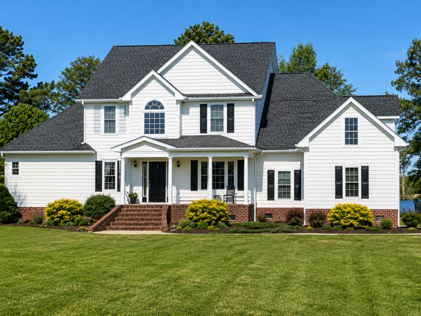 Large white two-story suburban house with black roof, brick porch, and green lawn under a clear blue sky