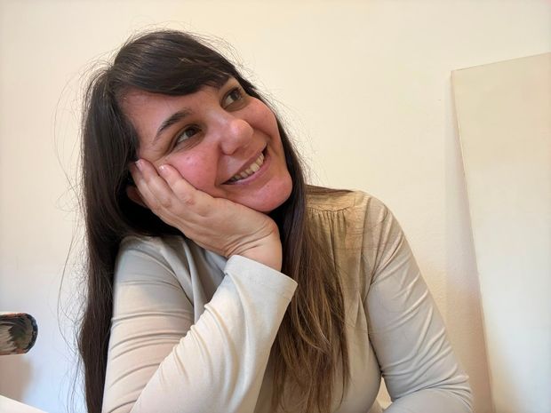Mujer sonriente con cabello largo y oscuro, apoyando el rostro en la mano, vestida con una blusa beige en el interior.
