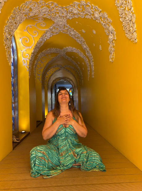 Una mujer vestida de verde está sentada en un pasillo con arco dorado, con las manos entrelazadas y una expresión serena.