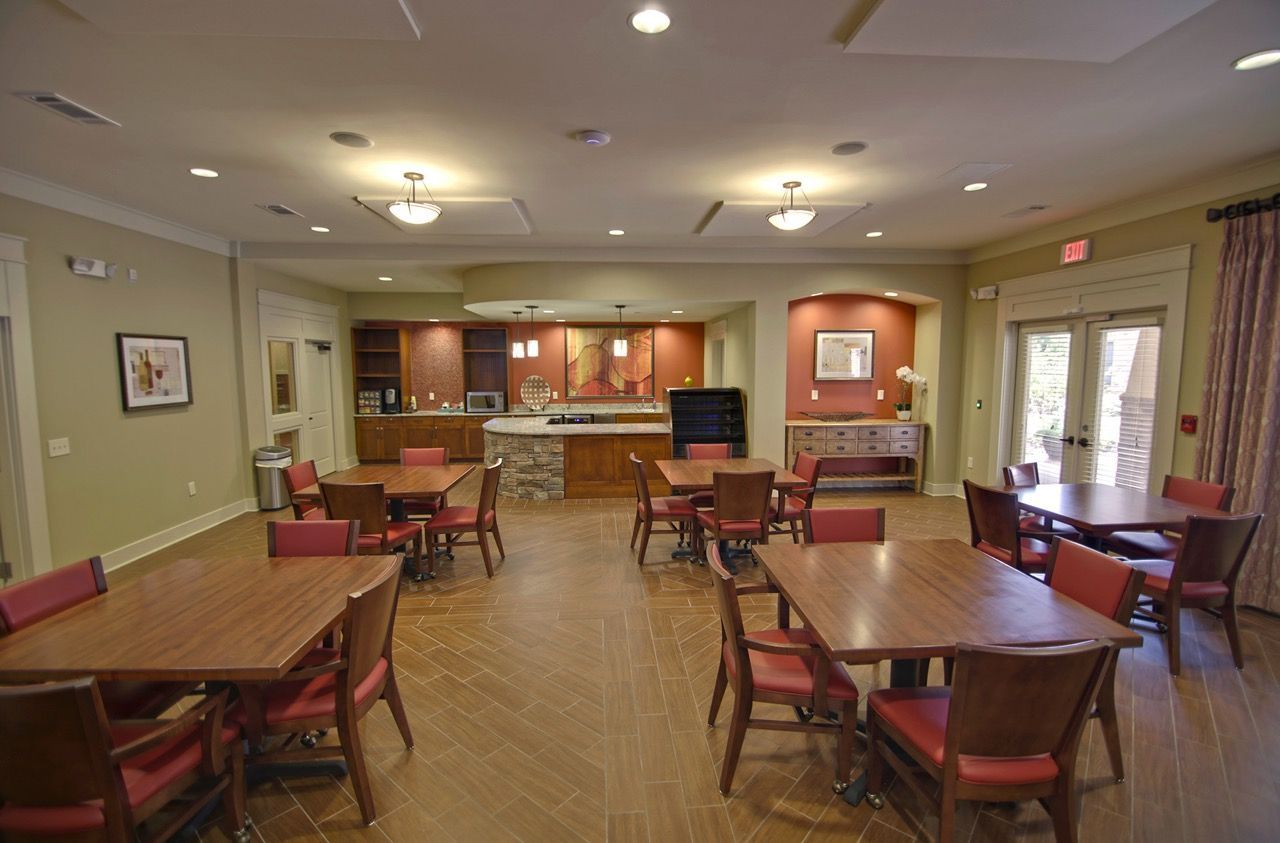 Interior of a resident community room with wooden tables, red chairs, and a kitchenette area.