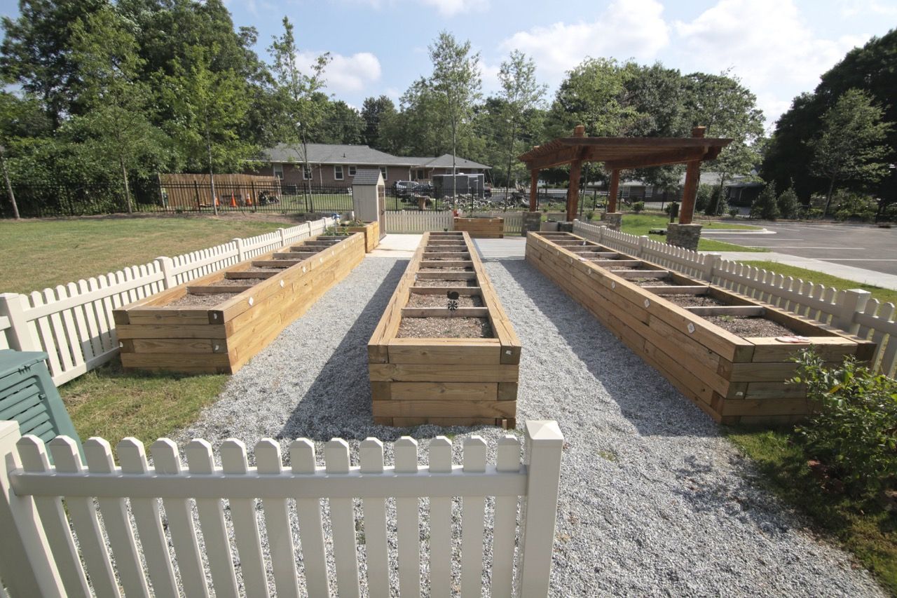 Community garden with four raised wooden planters, gravel paths, and a white picket fence.