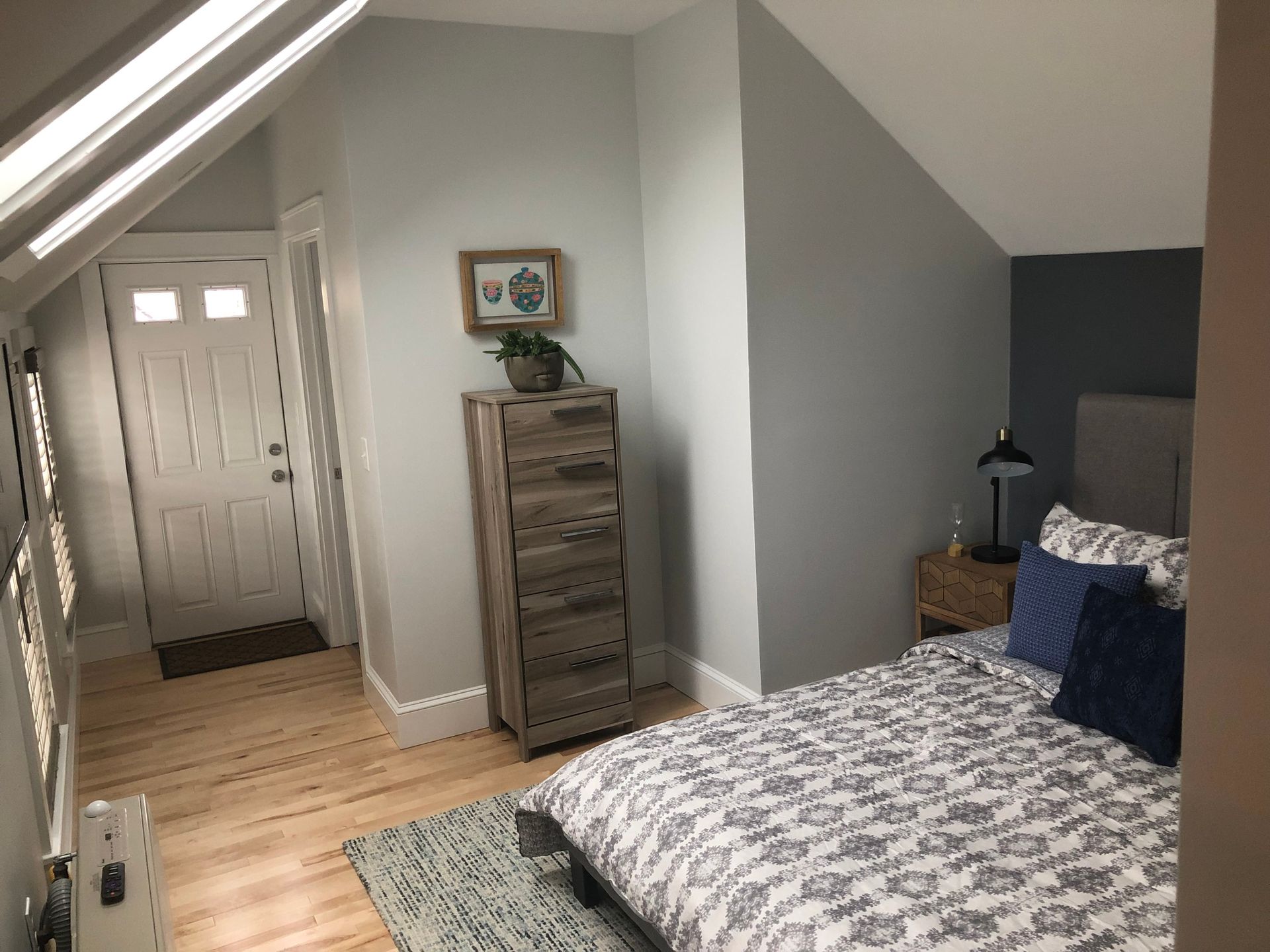 Bedroom with bed, dresser, and door. Light blue walls, wood floor, and natural light.
