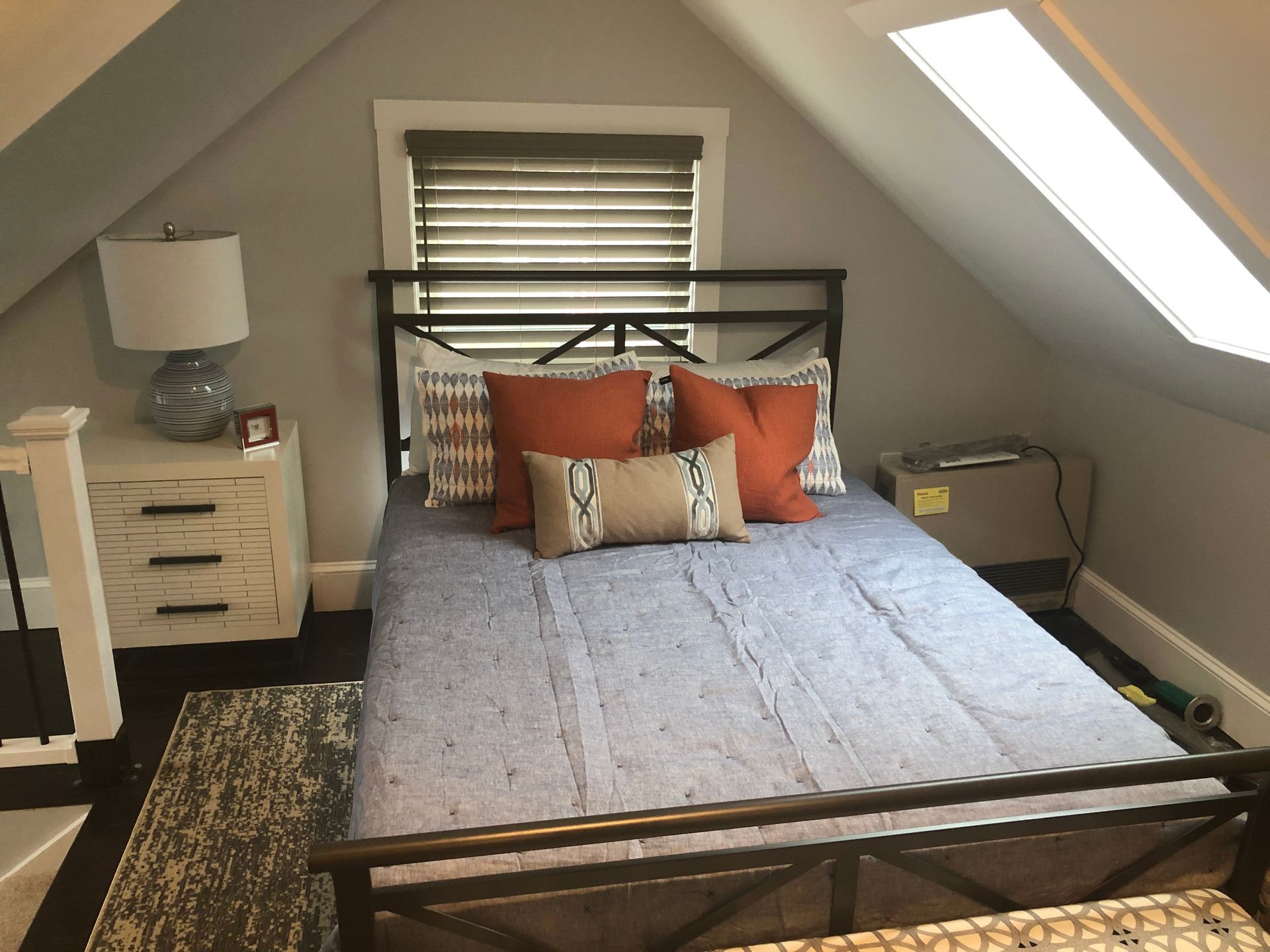 Bedroom with bed, nightstand, and window in a sloped-ceiling room. Blue bedding and orange pillows.