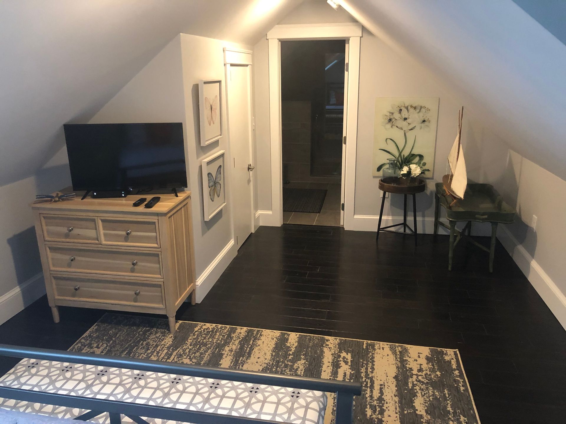 Bedroom with dark hardwood floors, dresser with TV, rug, small table with decor, and door to another room.