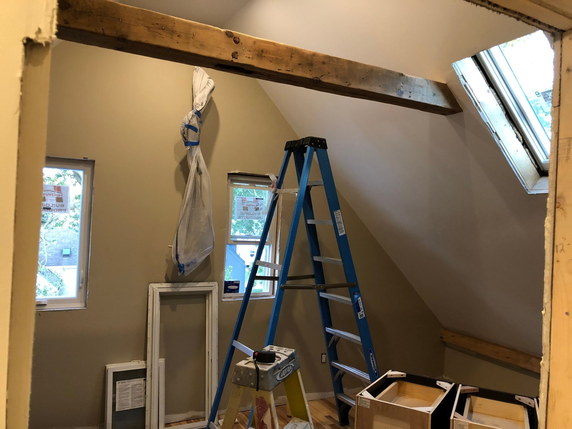 Construction in a room with a blue ladder, unfinished walls, wood beams, and small windows.