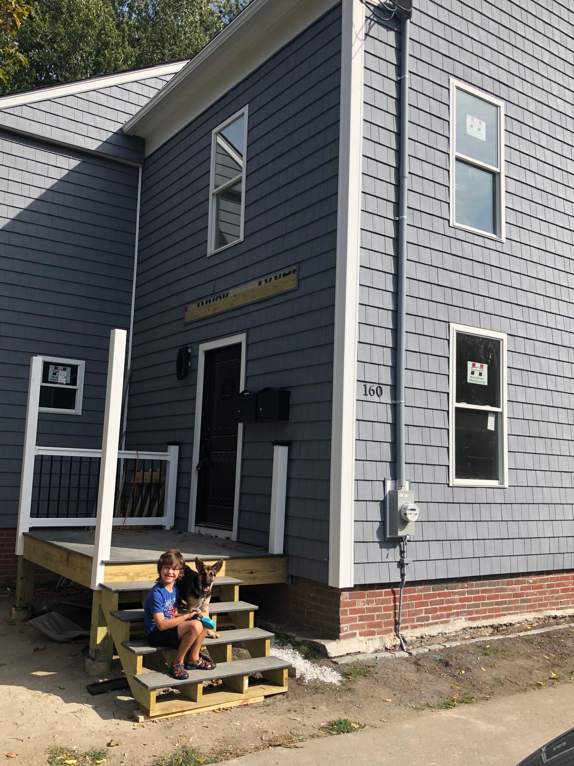 Boy sitting on steps of gray shingle house. Door and windows are visible.