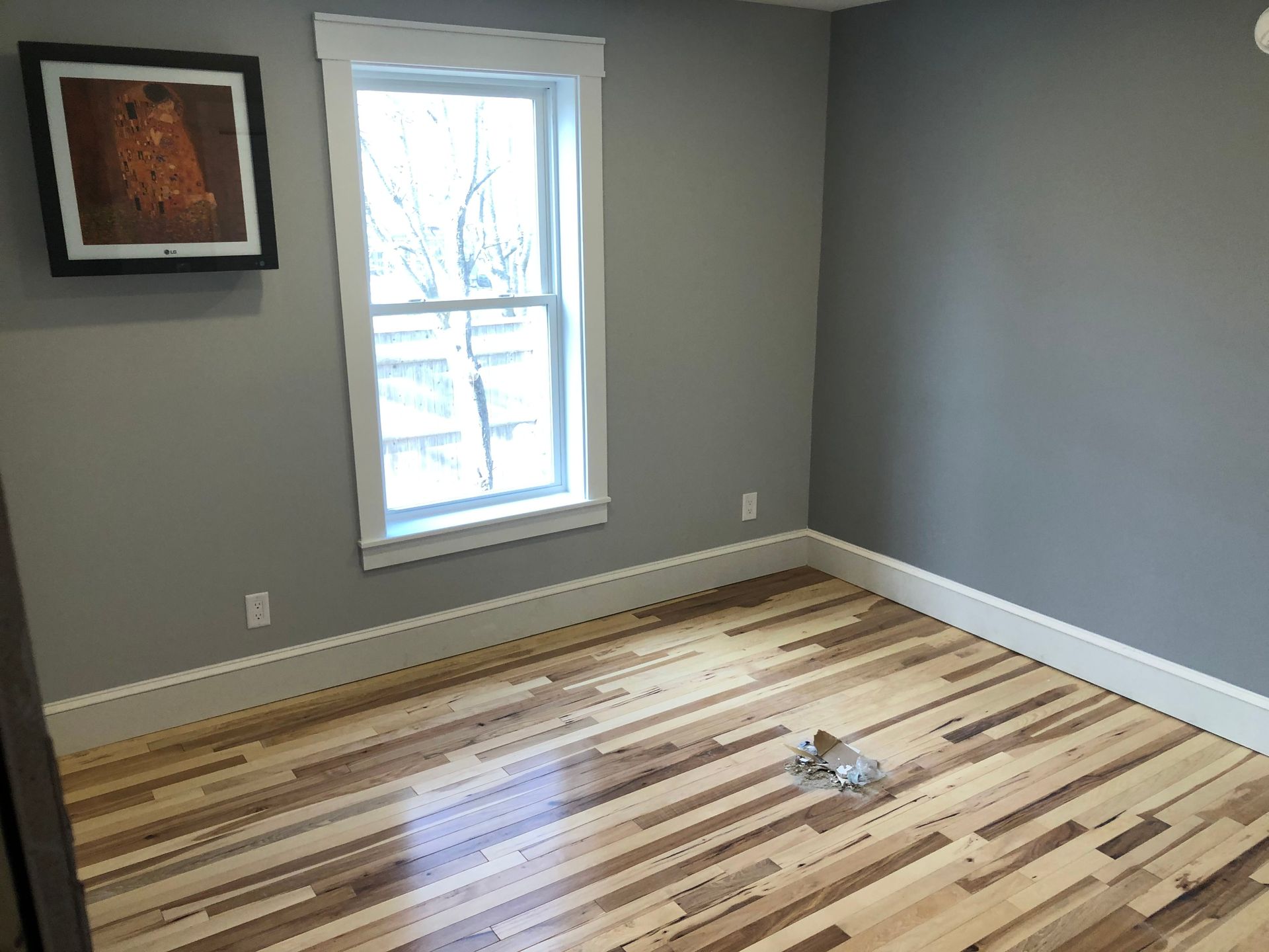 Empty room with gray walls, hardwood floor, window, and a framed painting.