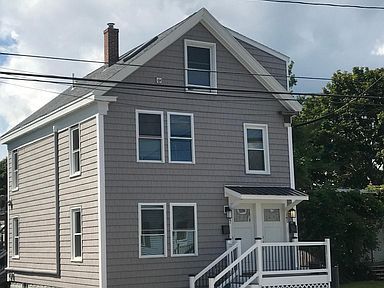 Gray two-story house with white trim, front porch, and windows. Overcast sky in the background.