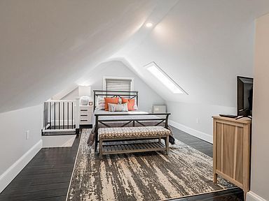 Bedroom in an attic with slanted ceilings, bed, rug, dark wood floor, white walls, and a small TV.