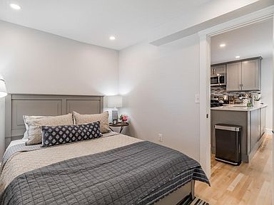 Bedroom with bed and side table, doorway to kitchen with cabinets and appliances.