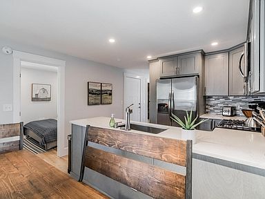 Modern kitchen with gray cabinets, stainless steel appliances, and a breakfast bar. A bedroom is visible in the background.