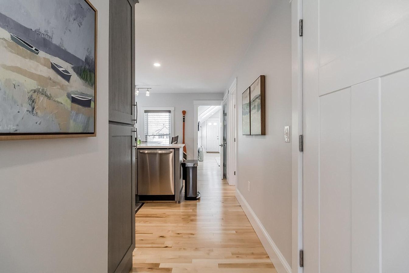 Hallway with light wood floors, leading to kitchen with stainless steel appliances. Painting on the left wall.