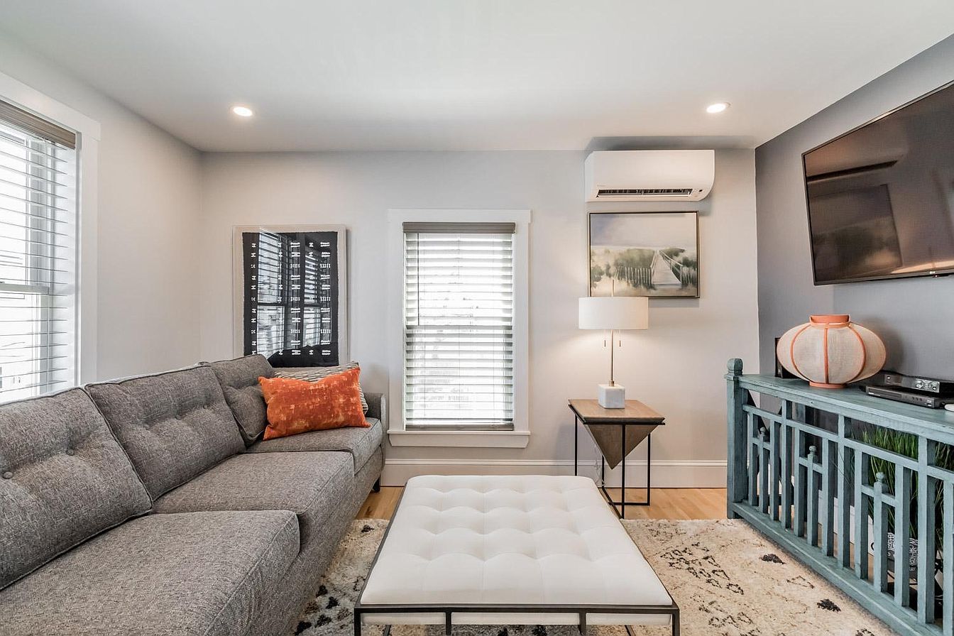 Living room with gray sofa, ottoman, and teal TV stand. Light walls, wood floors, and window blinds.