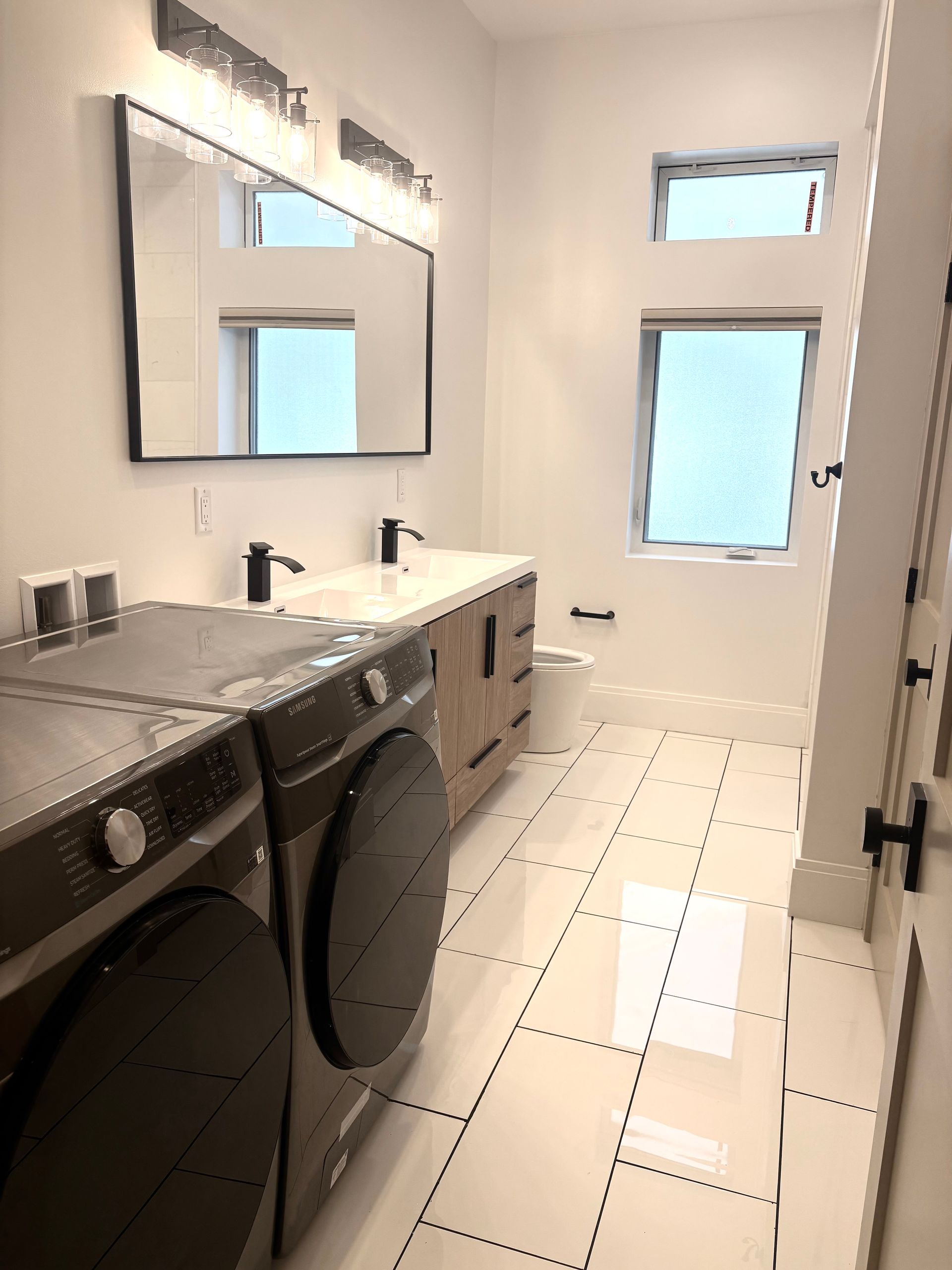 Laundry room with washer, dryer, vanity, mirror, two windows, and white tile floor.