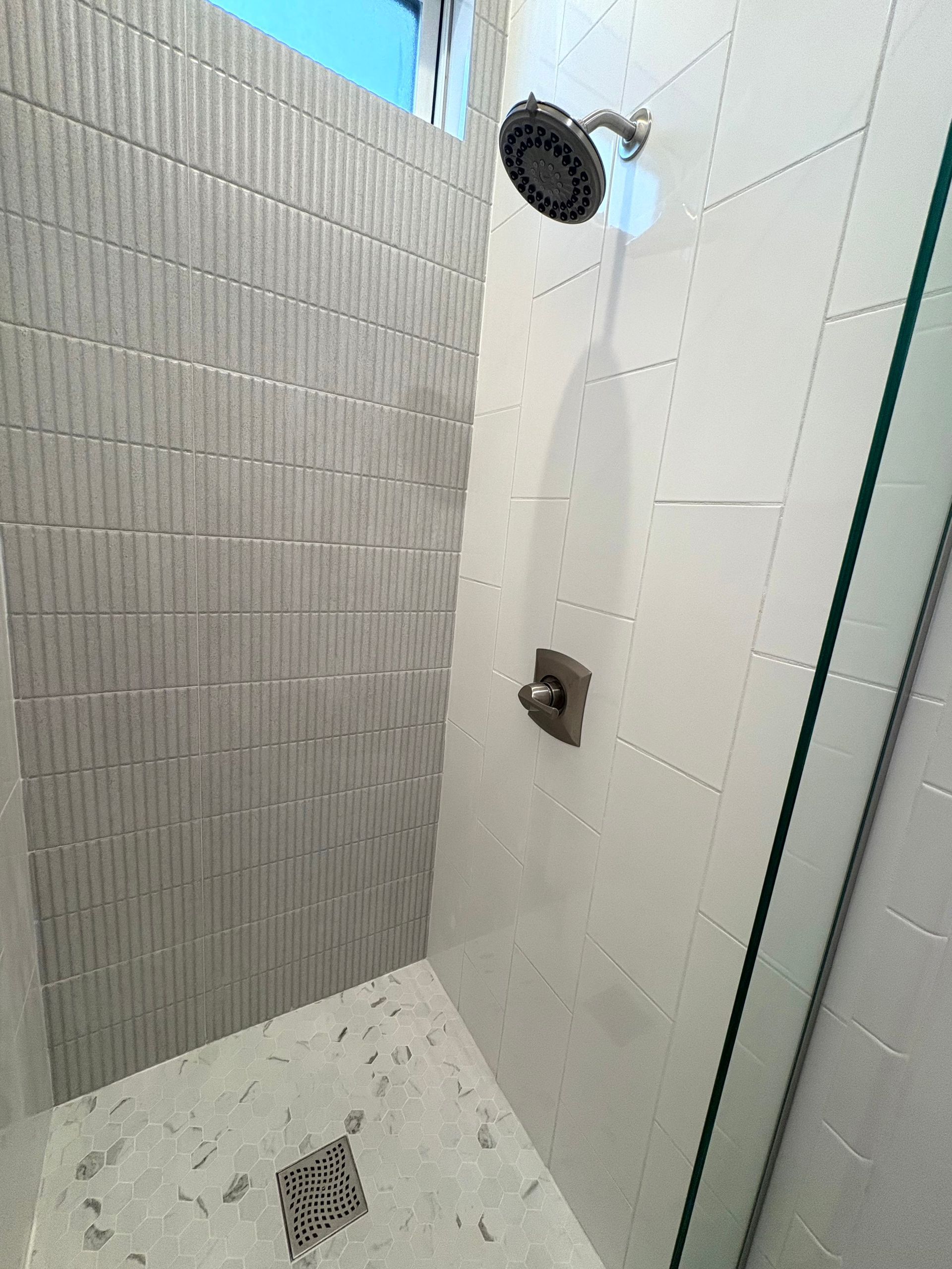 Shower with textured grey tile accent wall, white tile walls, glass door, and dark showerhead.