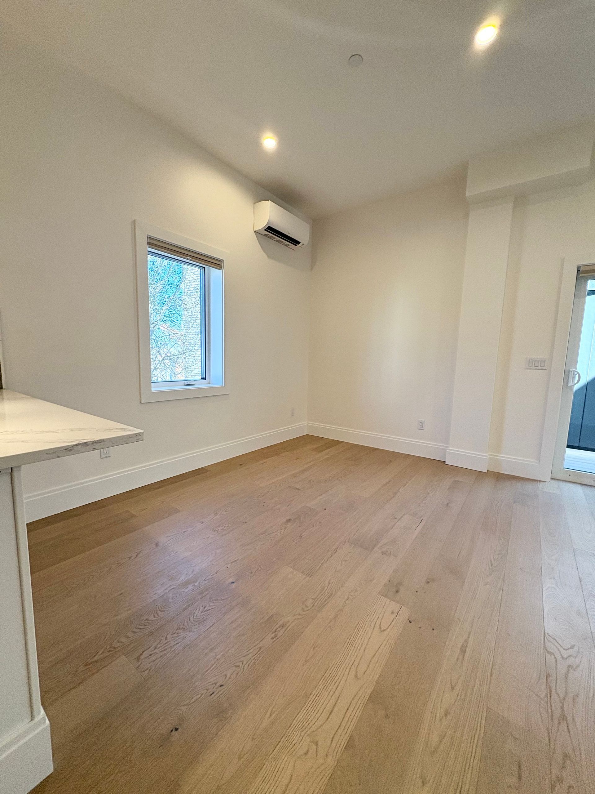 Empty room with hardwood floors, white walls, and a window; a mini-split air conditioner is mounted on the wall.