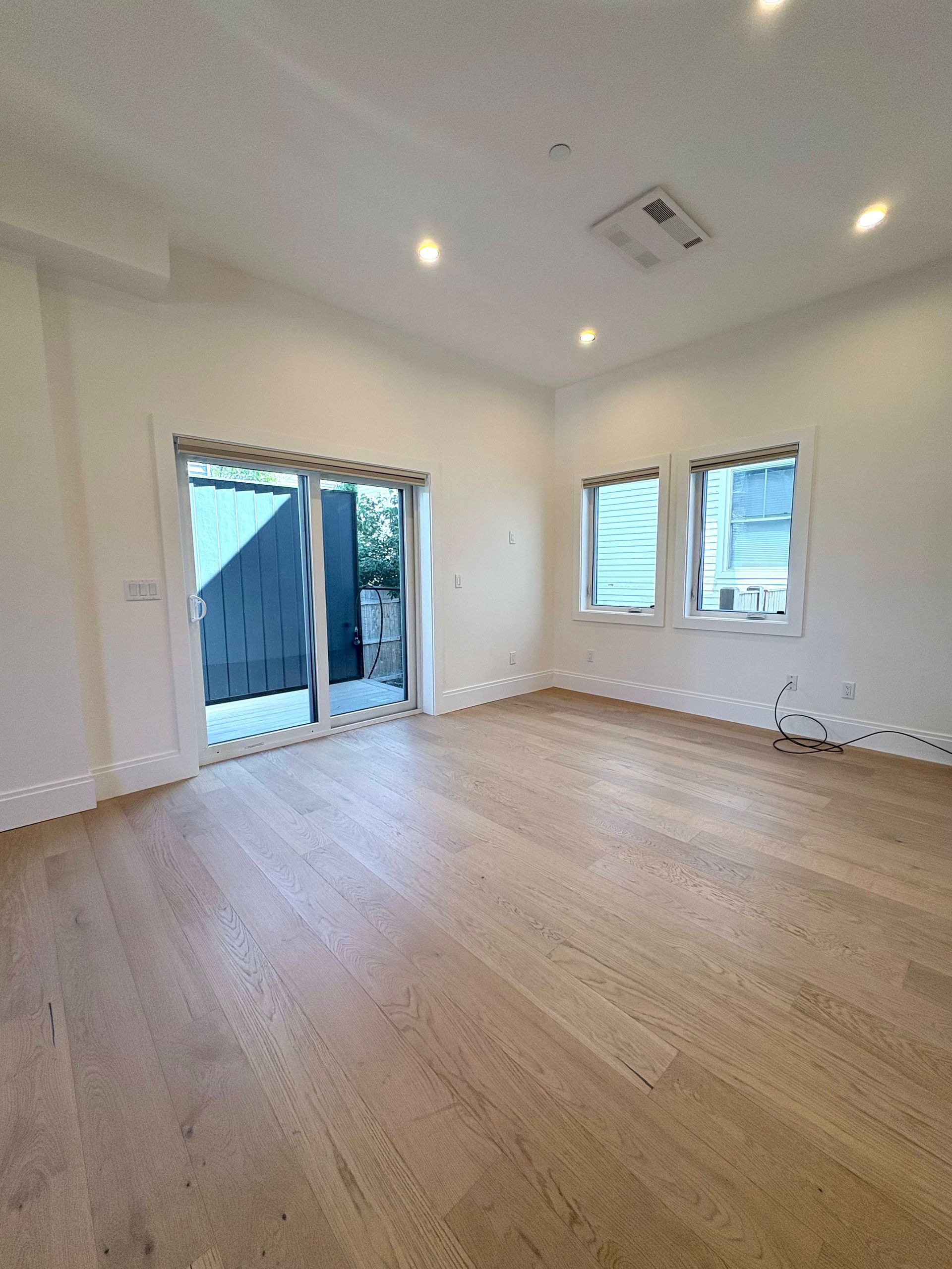 Empty, light-filled room with wood floor, sliding glass door, and two windows. White walls, recessed lighting.