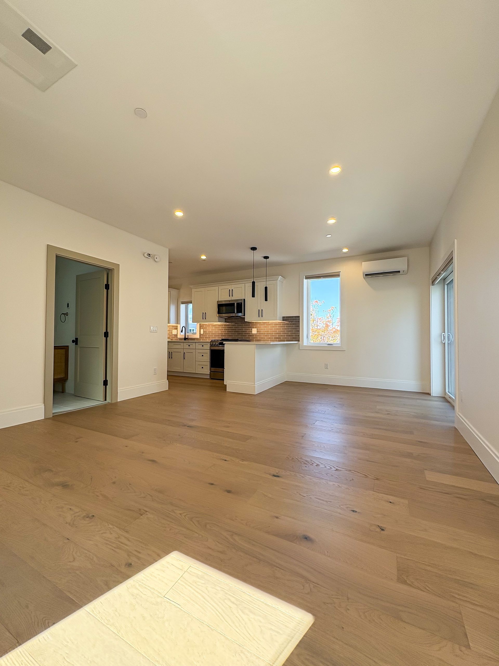 Empty, bright interior with hardwood floors. Kitchen visible in the back, open floor plan.