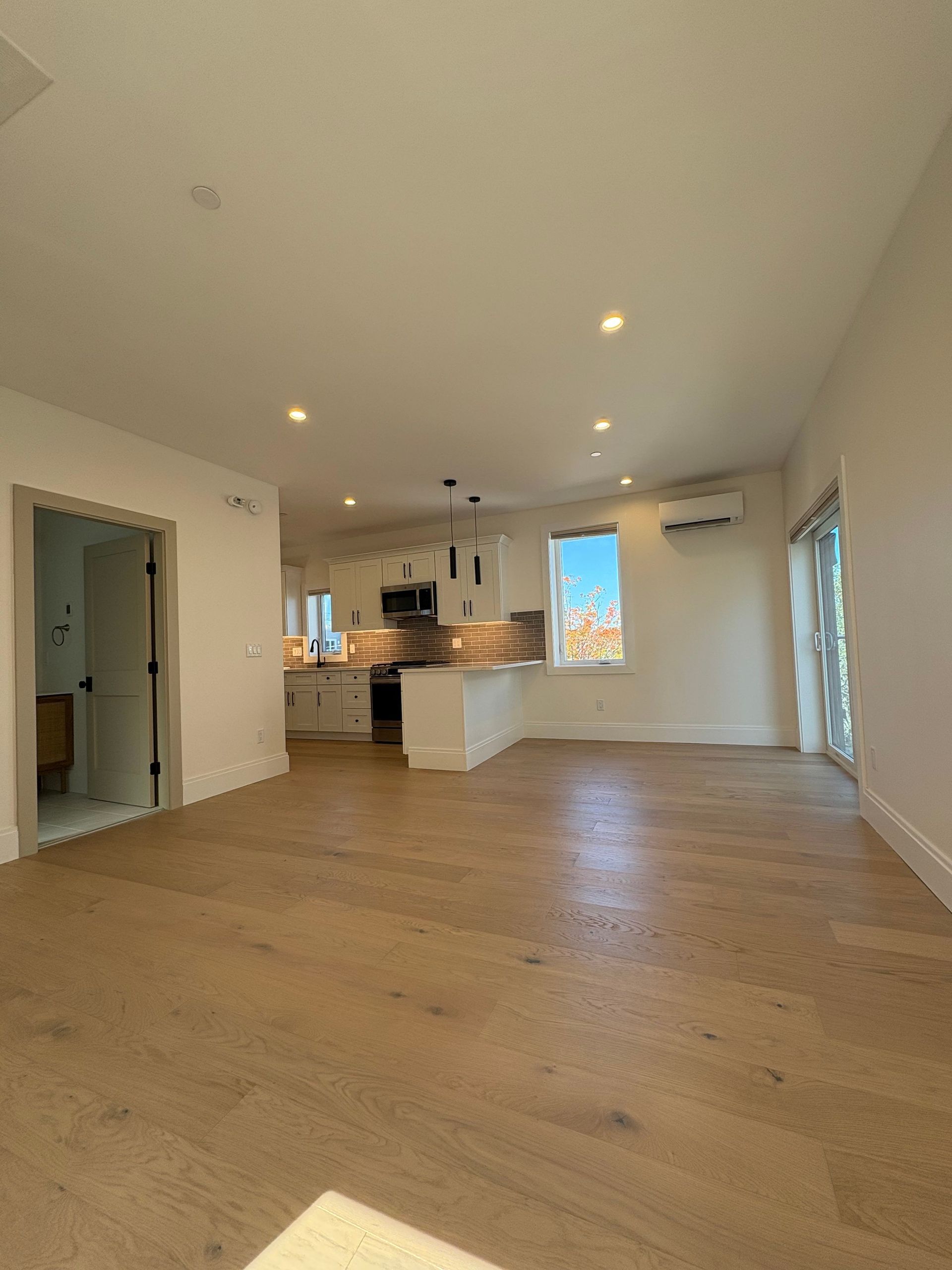 Empty, bright living room with hardwood floors, a kitchen, and a sliding door.
