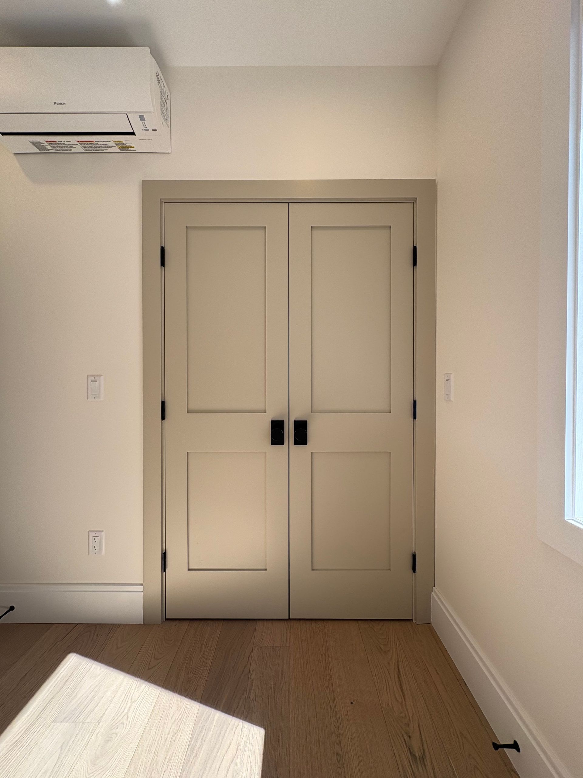 Closed beige double doors, black hardware, inside a room with light wood floors and white walls.