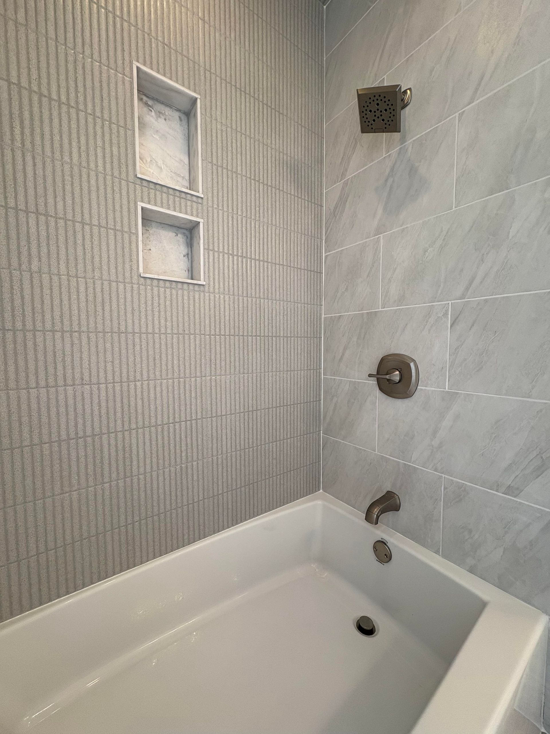 Gray-tiled bathroom with white tub. Two built-in shelves on textured wall. Showerhead and faucet are brushed nickel.