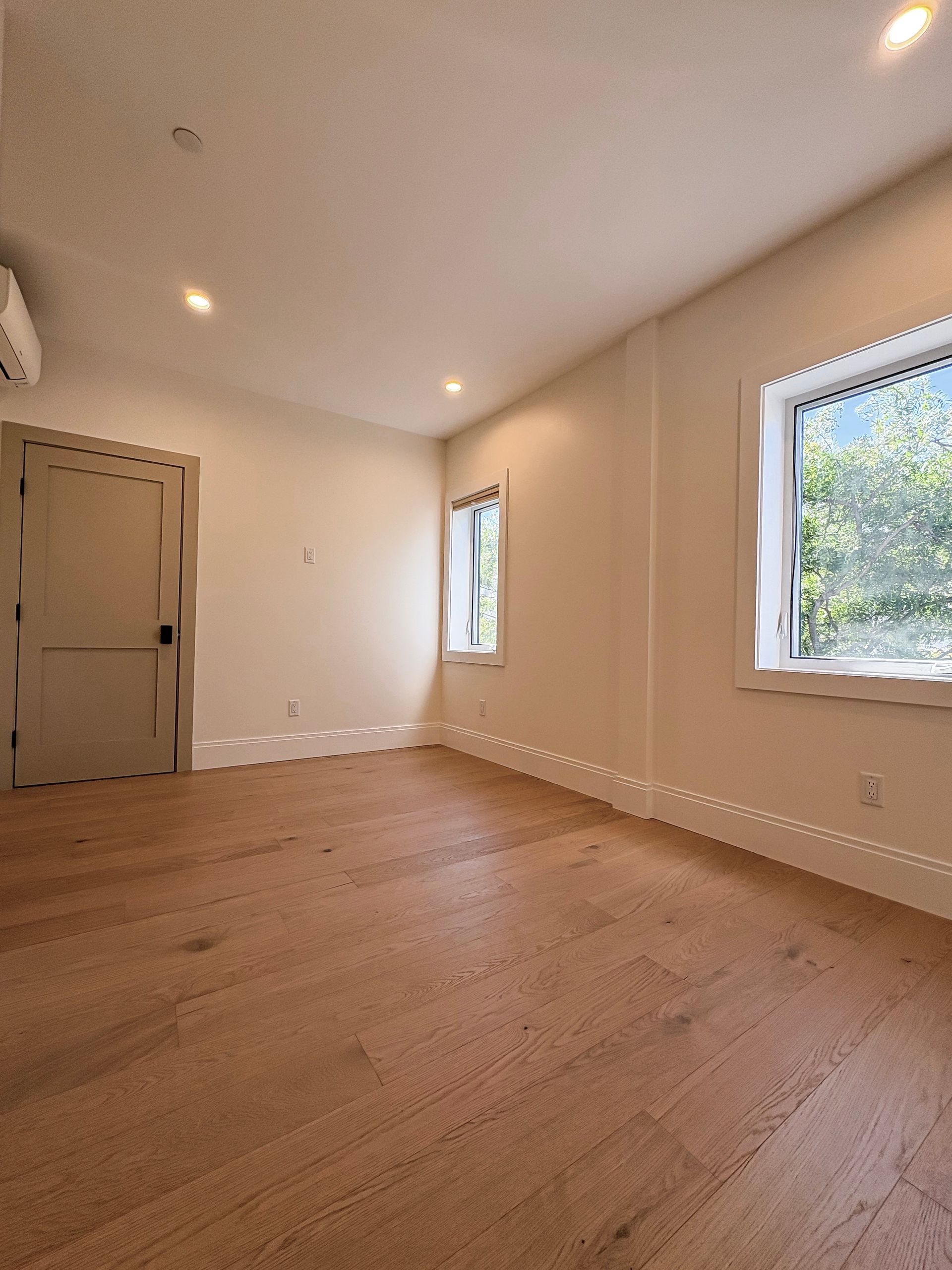 Empty room with wooden floor, two windows, and a door; walls are white.