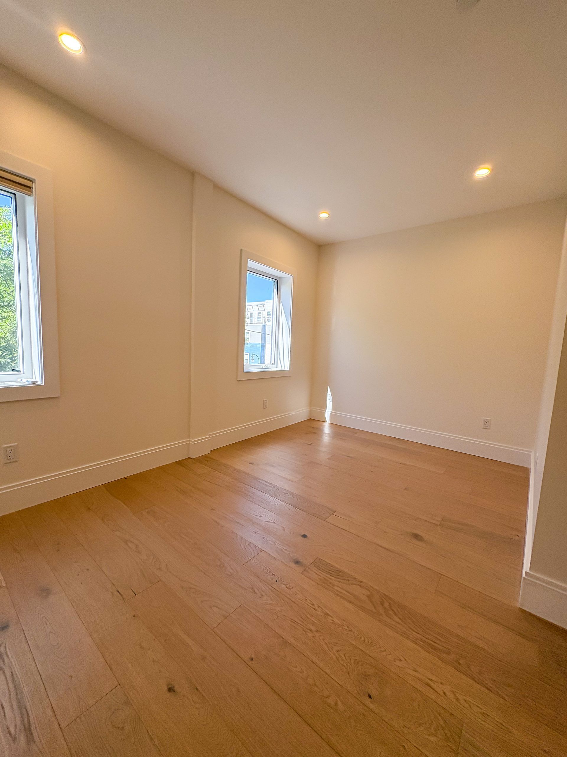 Empty room with wooden floor, two windows, and recessed lighting; beige walls.