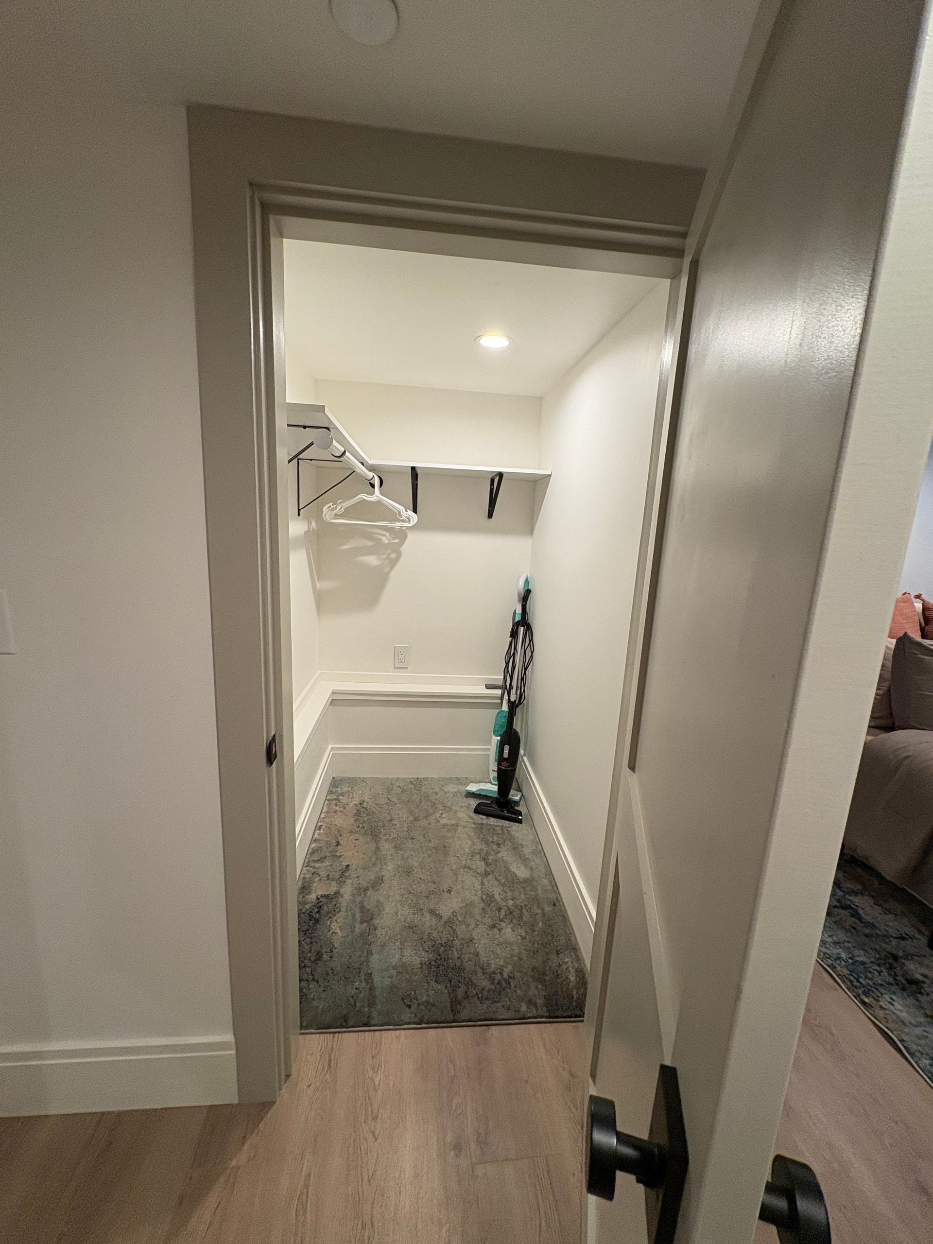 A narrow, white closet with shelves, a vacuum, and a grey rug; doorway visible.