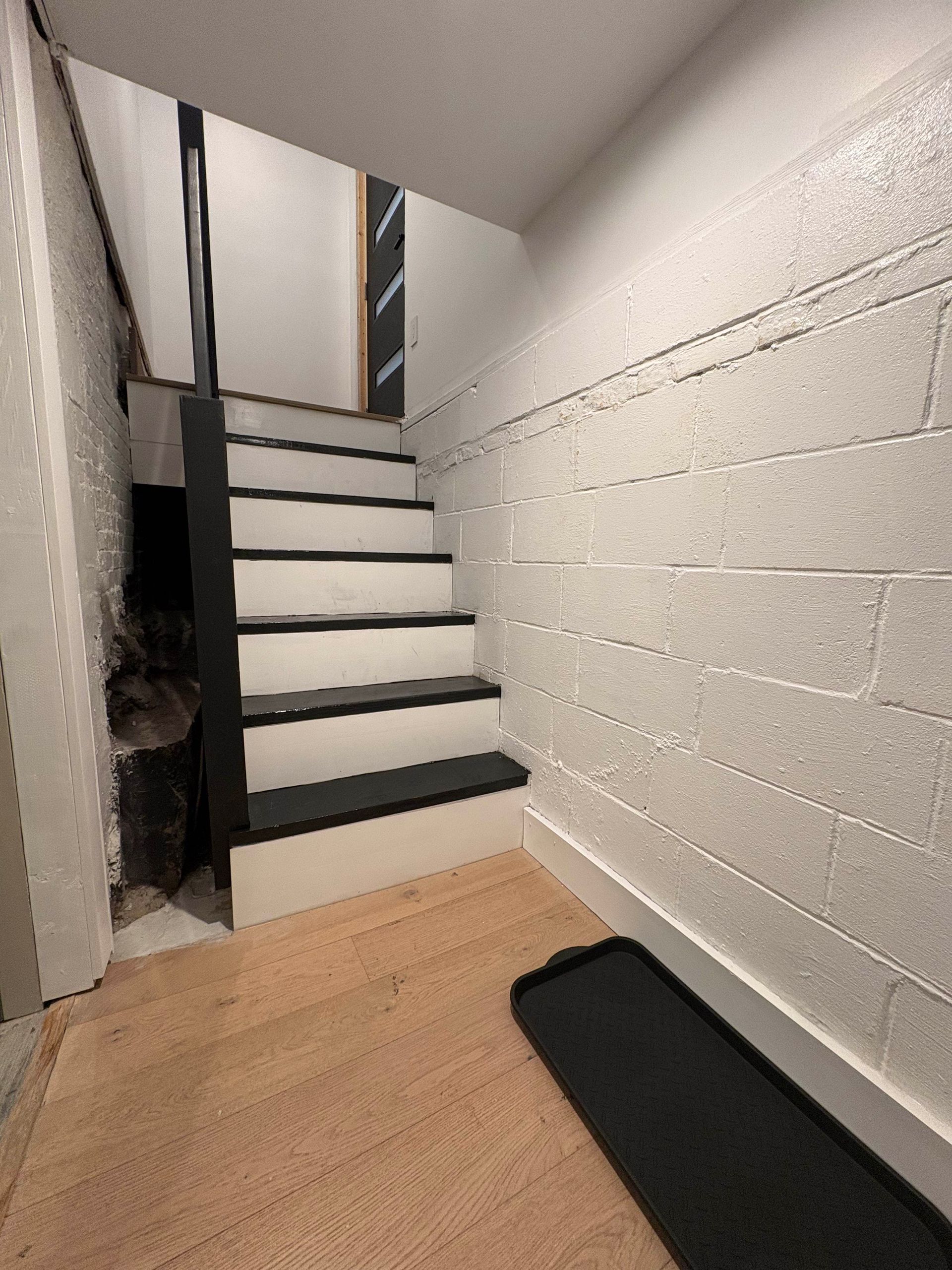 Staircase leading down, black and white steps, cinder block wall, hardwood floor.