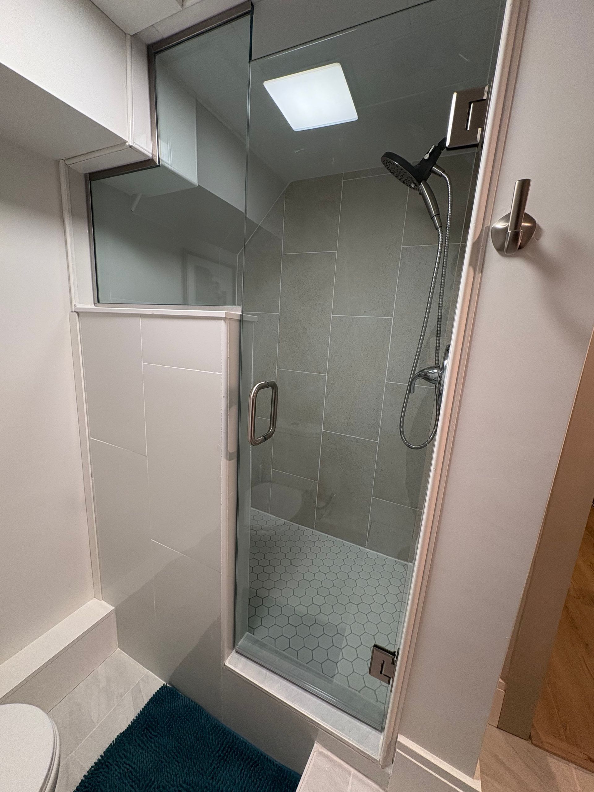 Shower stall with glass door and rain showerhead; white and gray tile; teal rug.
