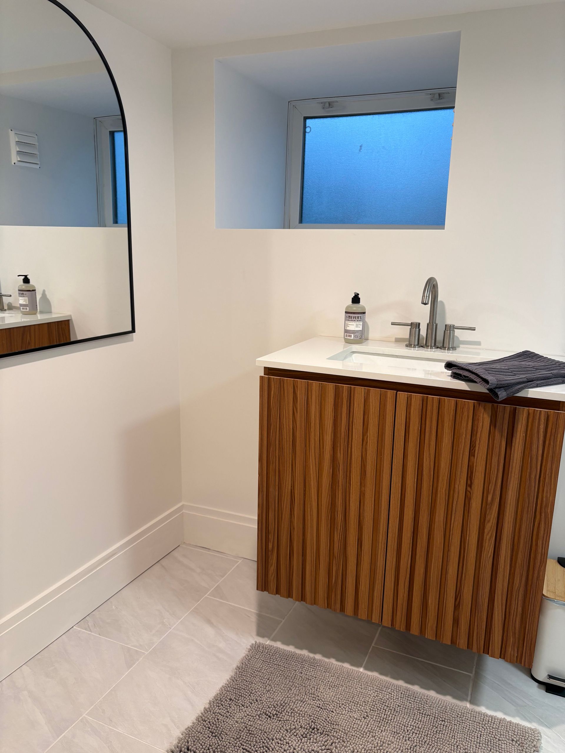 Bathroom with wooden vanity, arch mirror, window, white walls, and gray rug.
