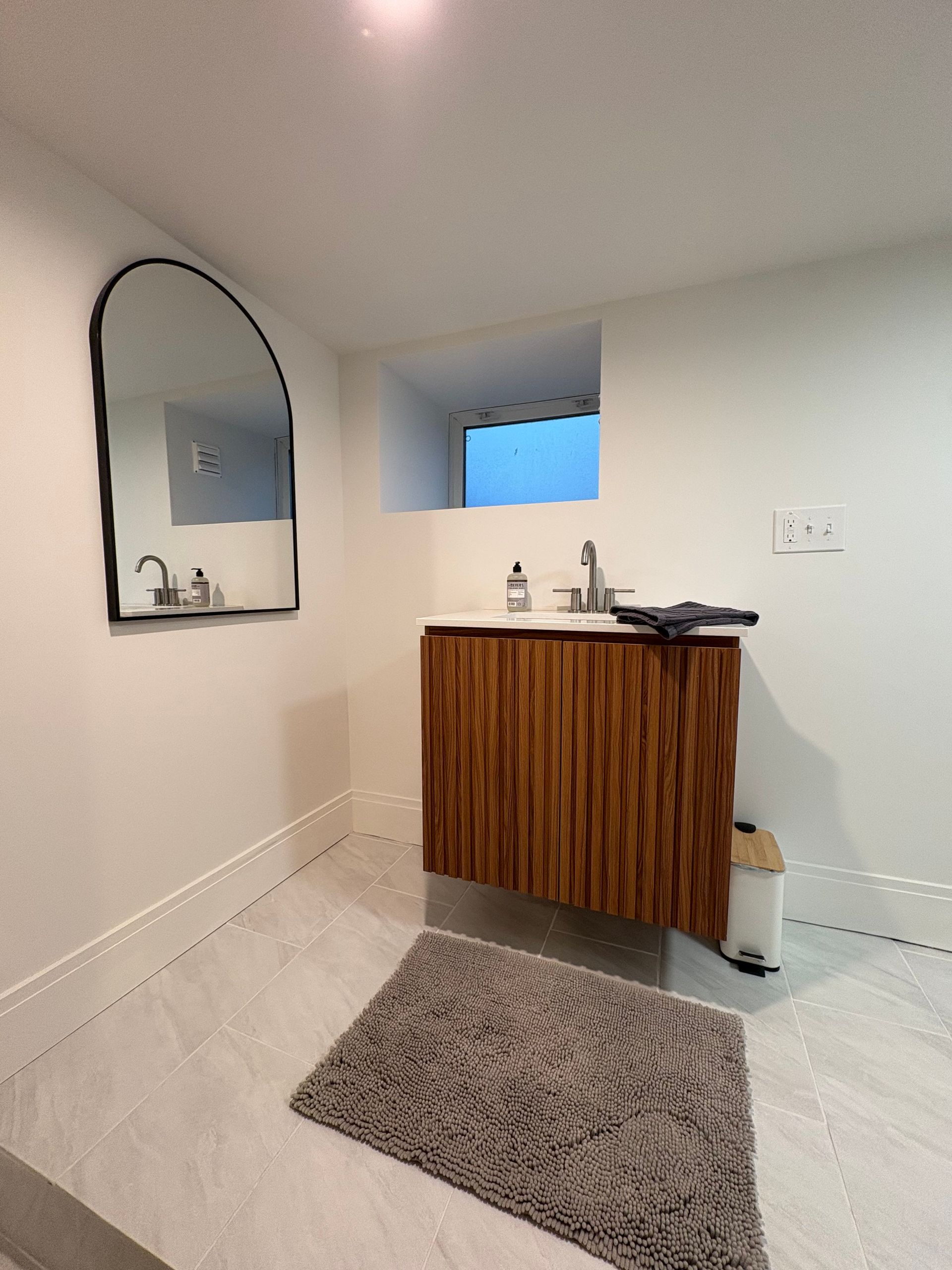 Bathroom with arched mirror, wooden vanity, and gray rug. Light-colored walls and floor.