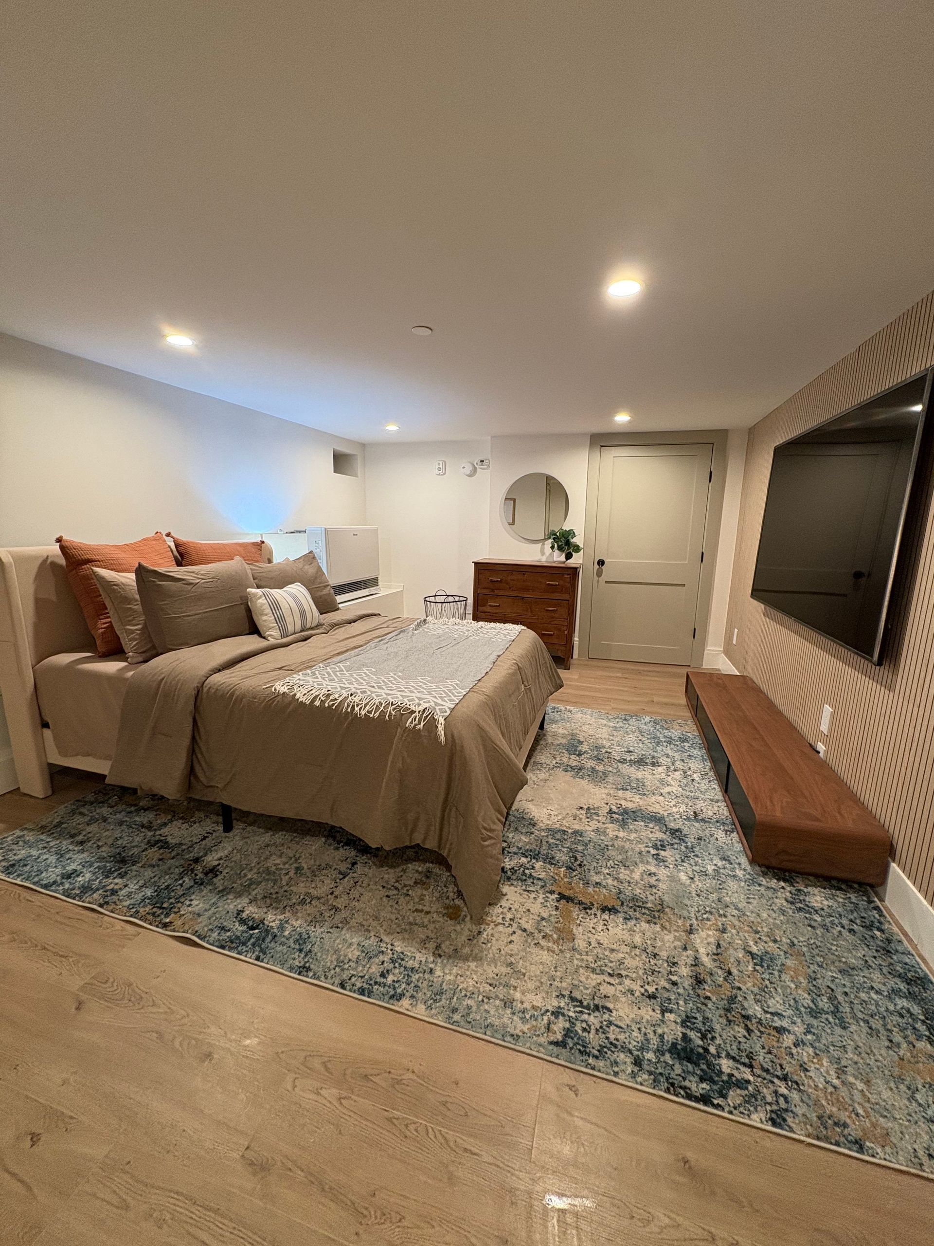 Bedroom with bed, rug, TV on patterned wall, dresser, and door. Blue and brown tones.