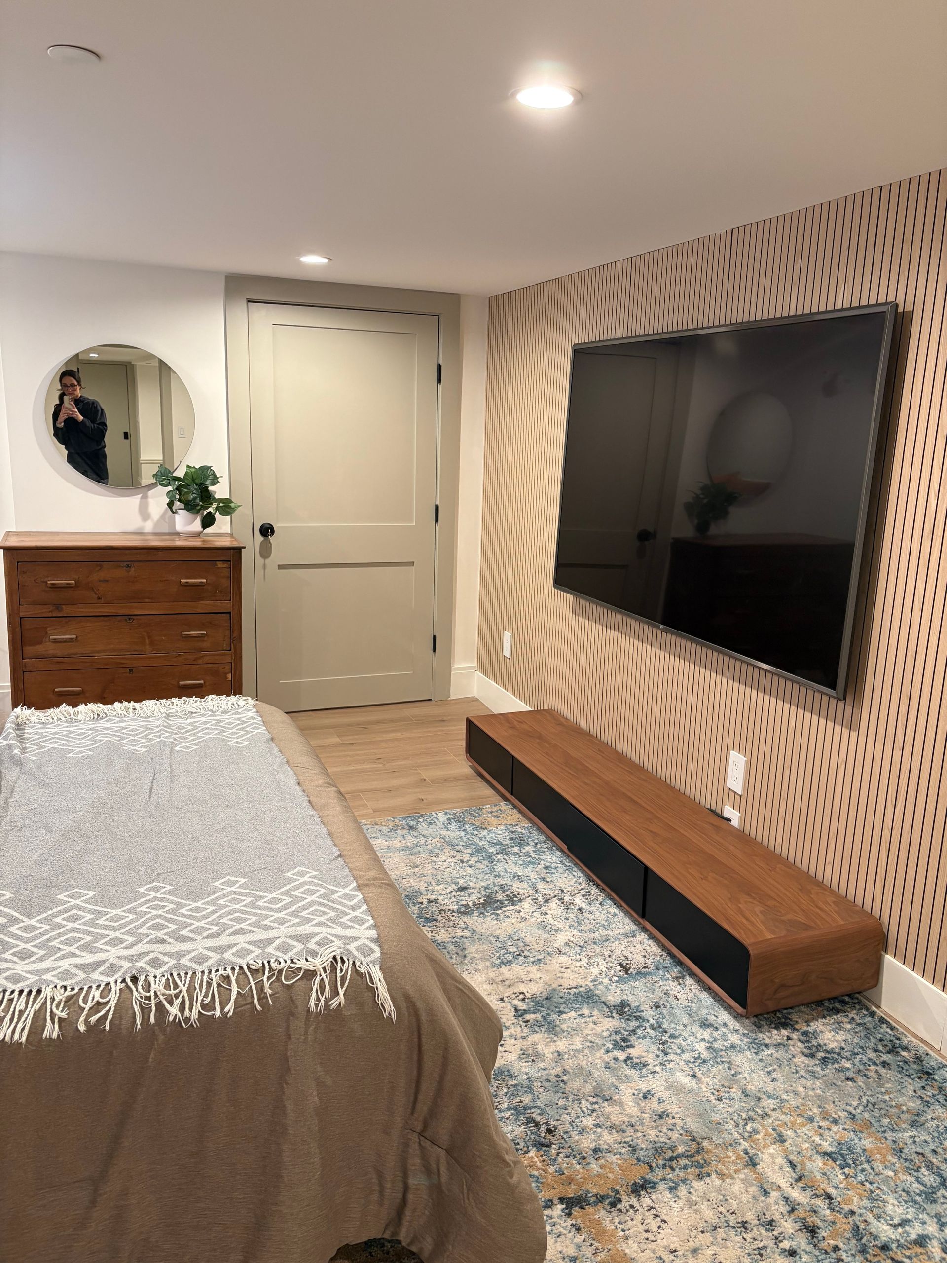 Bedroom with wall-mounted TV, wood console, patterned accent wall, and wood dresser. Blue rug and beige bed.