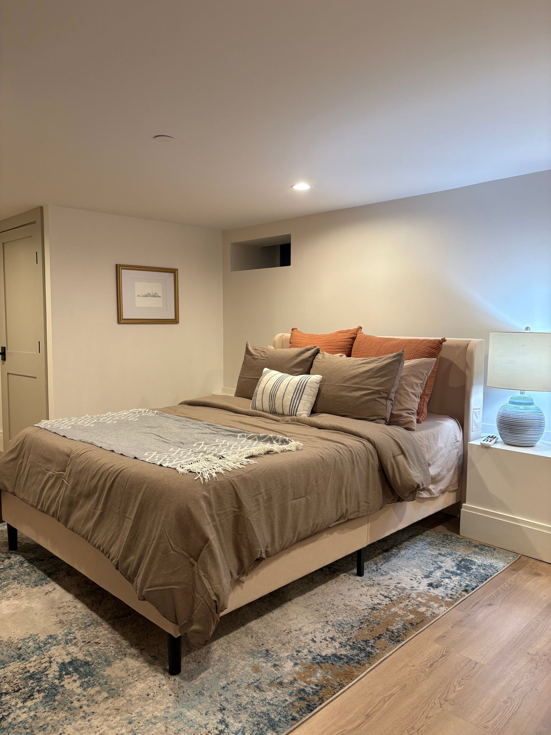 Bedroom with a bed, pillows, rug, and bedside lamp. Warm neutral colors.