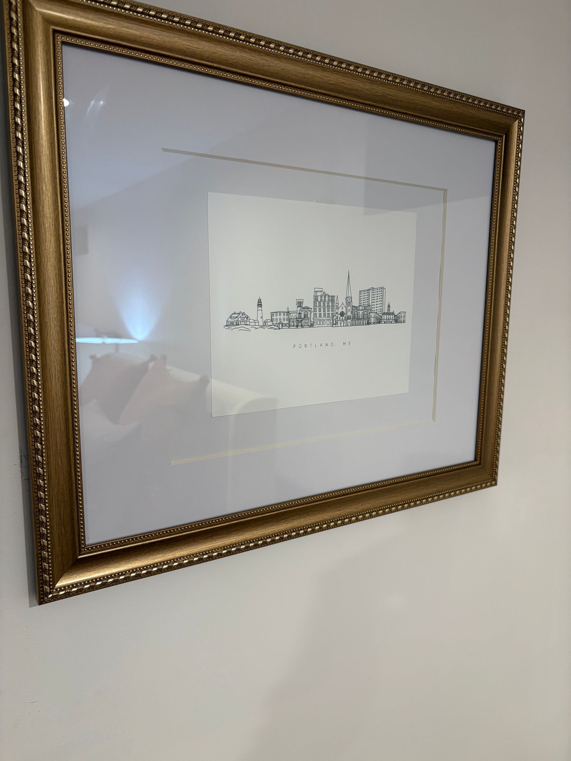 Gold-framed art piece on a white wall. The artwork is a simple, black and white sketch of a city skyline.