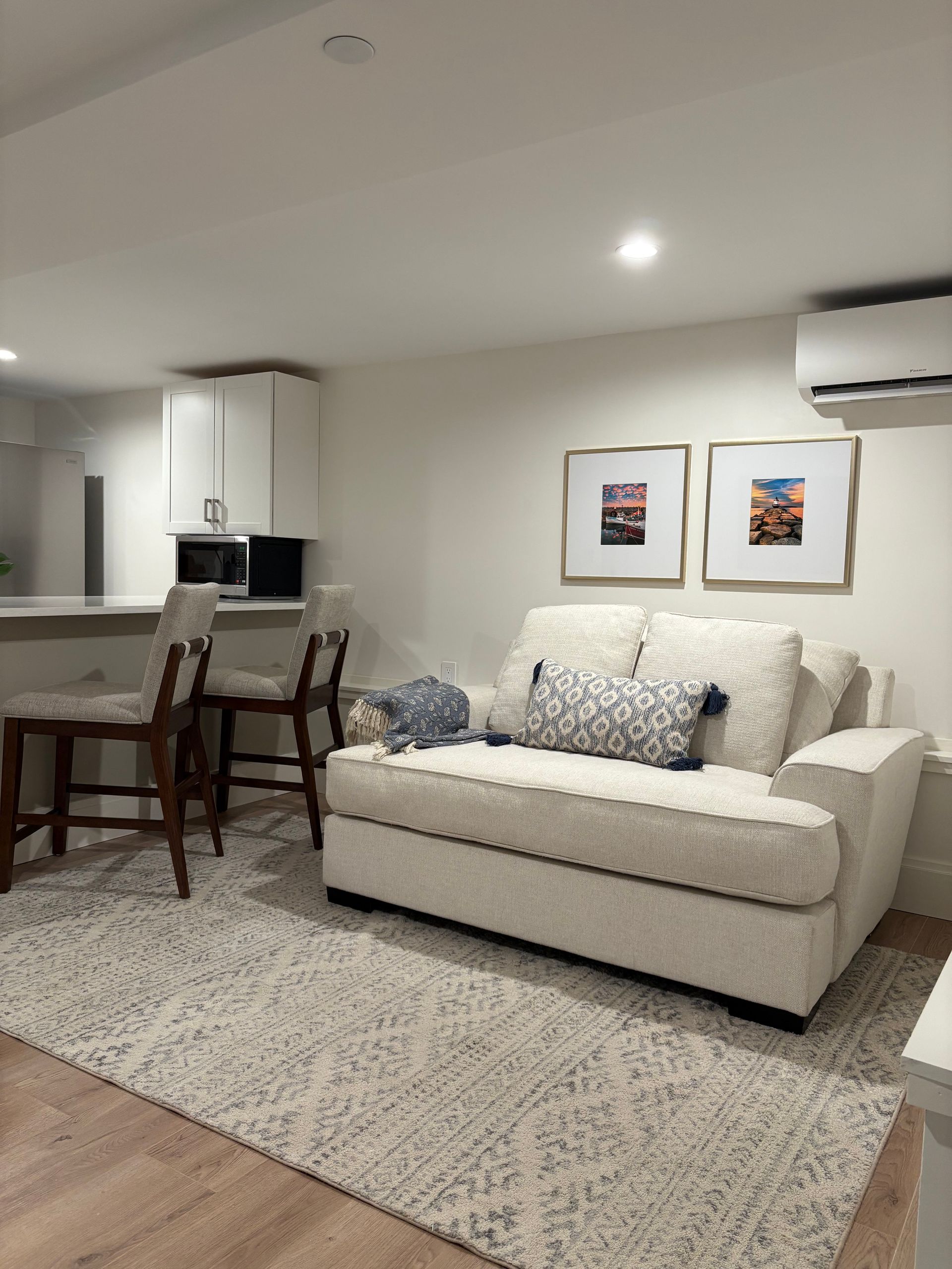 Cozy living space with white couch, rug, small kitchen with barstools, two framed pictures, and air conditioning unit.