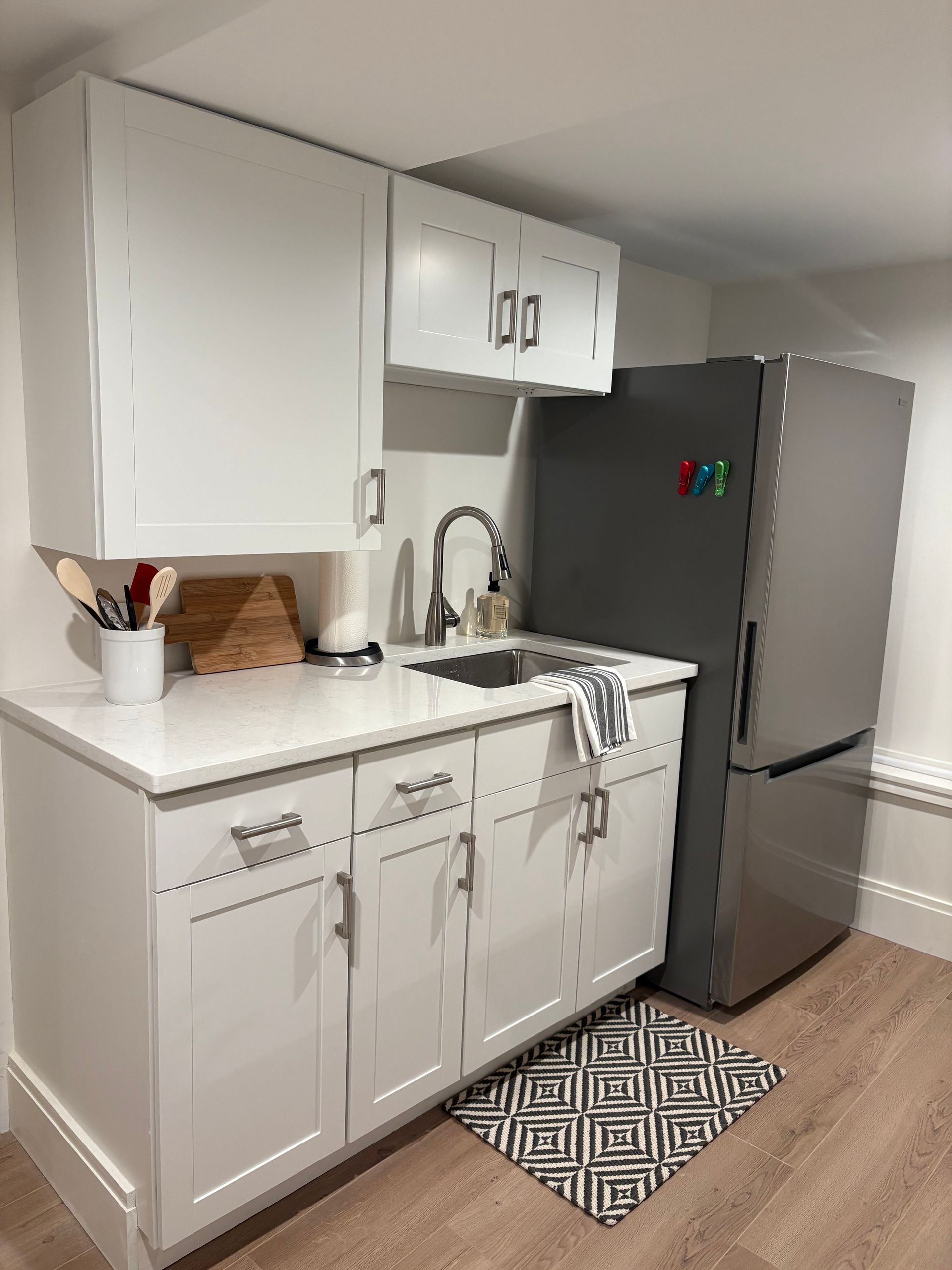 Small white kitchen with cabinets, sink, and refrigerator. Wooden floor and decorative rug.