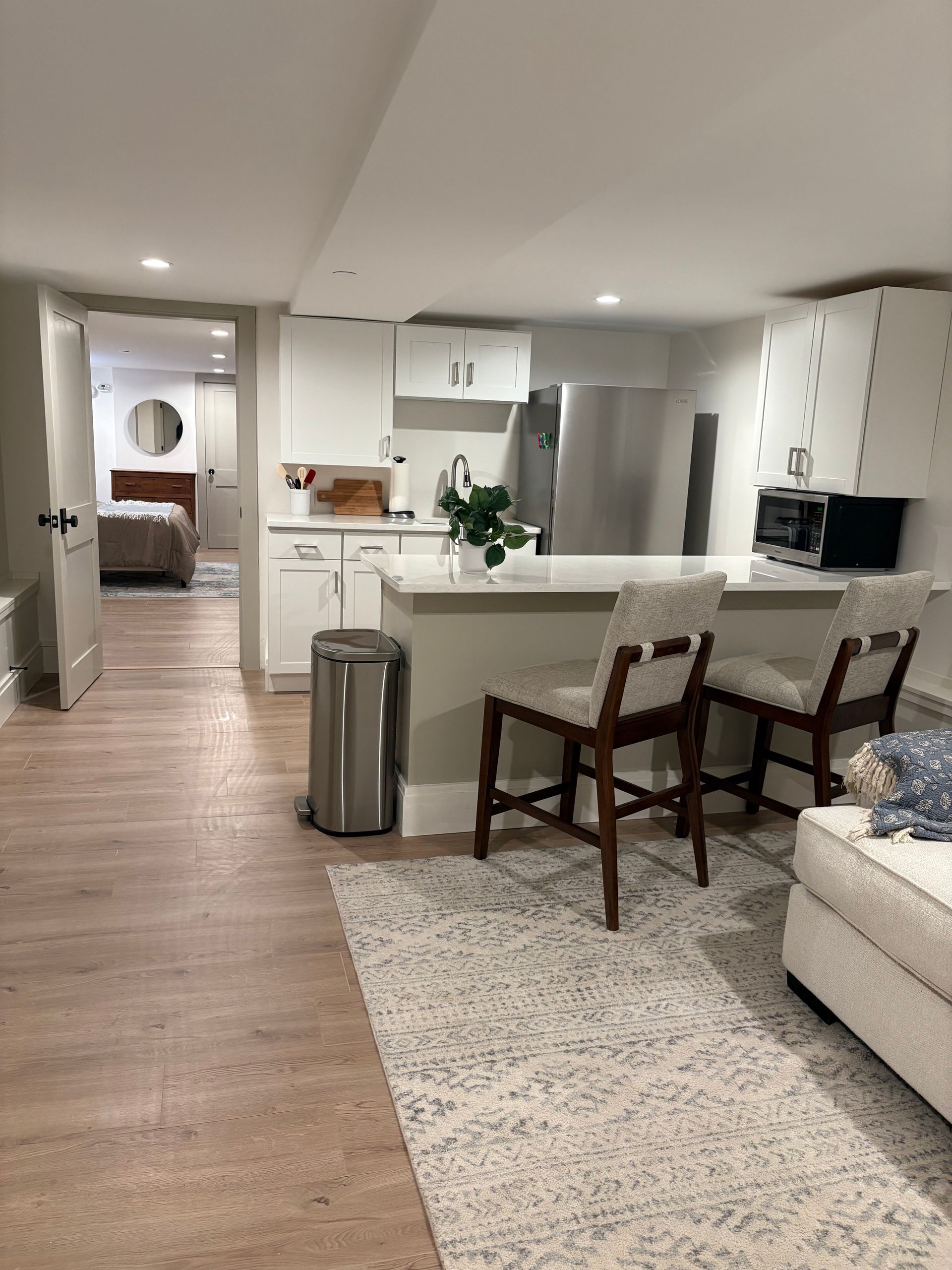 Kitchen area with white cabinets, stainless steel appliances, and a breakfast bar with two chairs, leading to a bedroom.