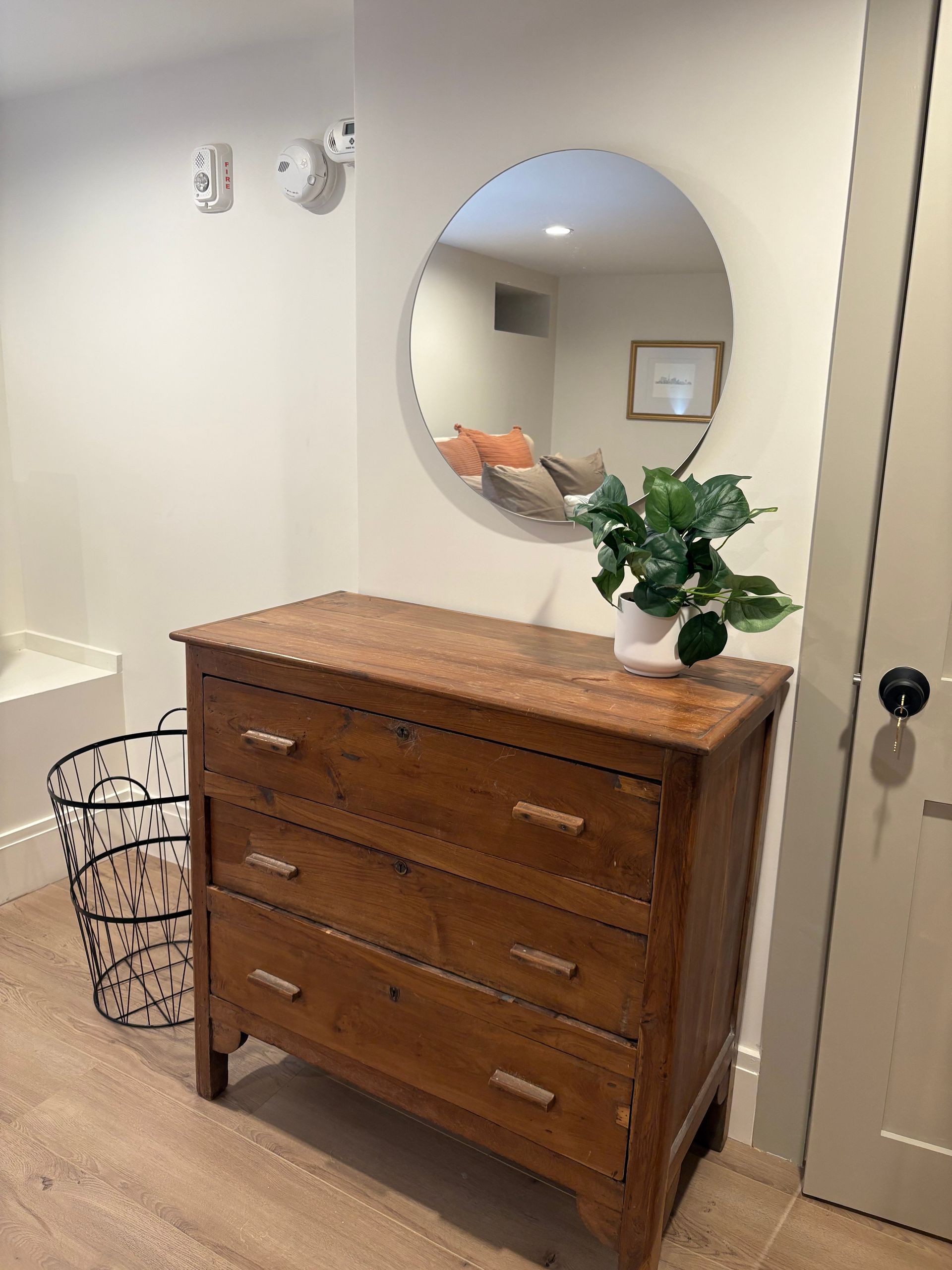 Wooden dresser with a round mirror and a plant. A wire basket sits beside it.