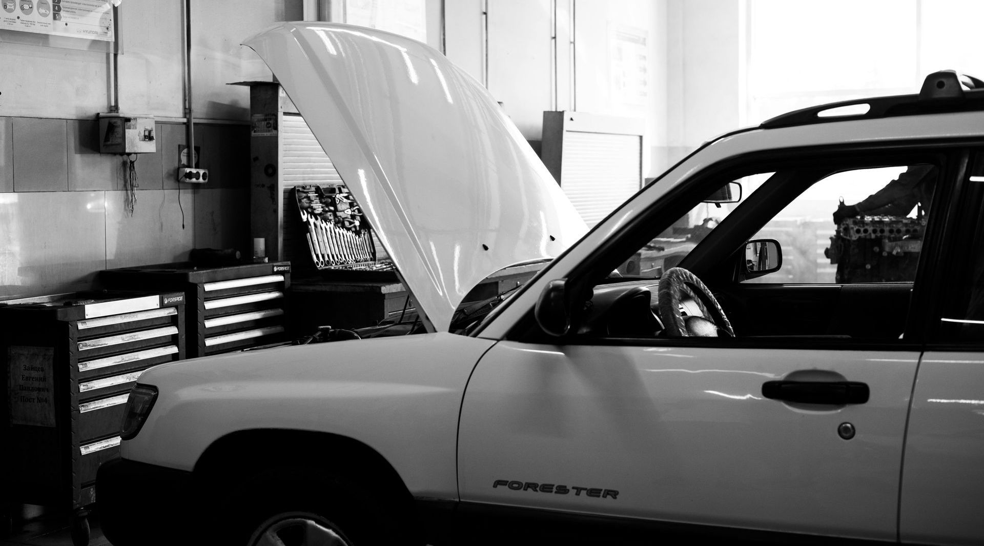 White car with hood open in a garage, tool chests visible.
